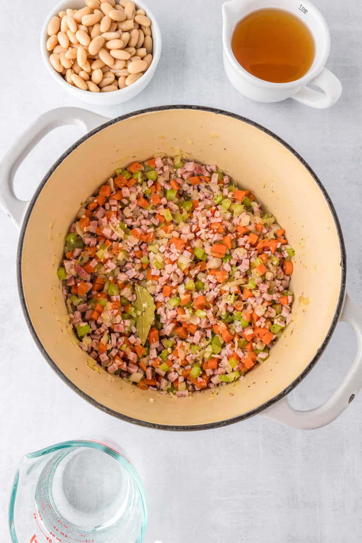 A pot containing diced ham, carrots, celery, and onion is being sautéed for white bean and ham soup; nearby are bowls of white beans, broth, and water.