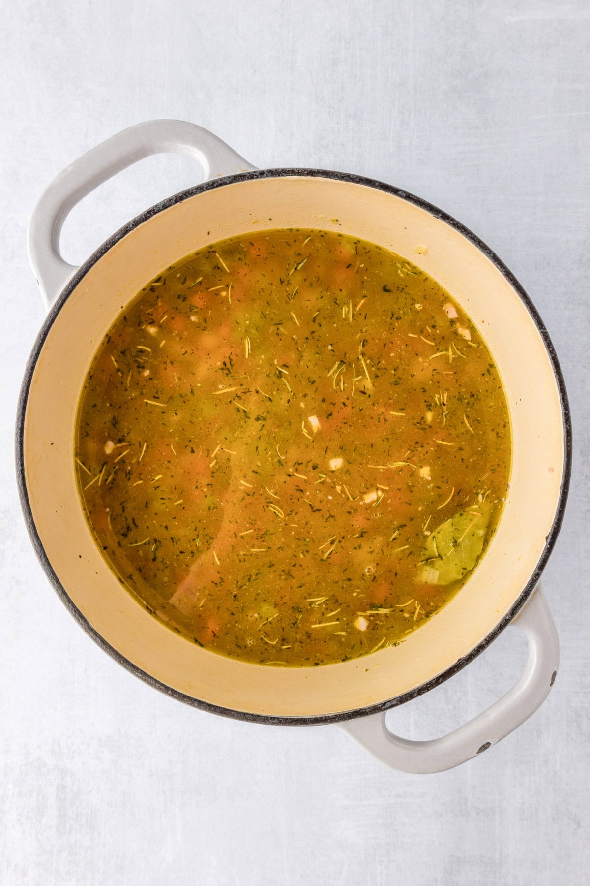 A white Dutch oven filled with herb-seasoned broth, vegetable pieces, and savory white bean and ham soup, set on a light-colored surface.