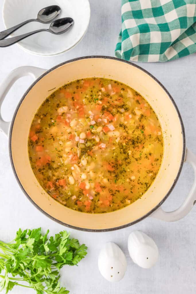 A white pot filled with hearty rosemary white bean and ham soup sits on a counter.