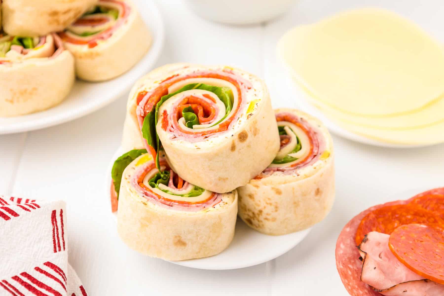 Three Italian pinwheels filled with deli meats, cheese, and lettuce are stacked on a white plate, with more pinwheels and sandwich ingredients nearby.