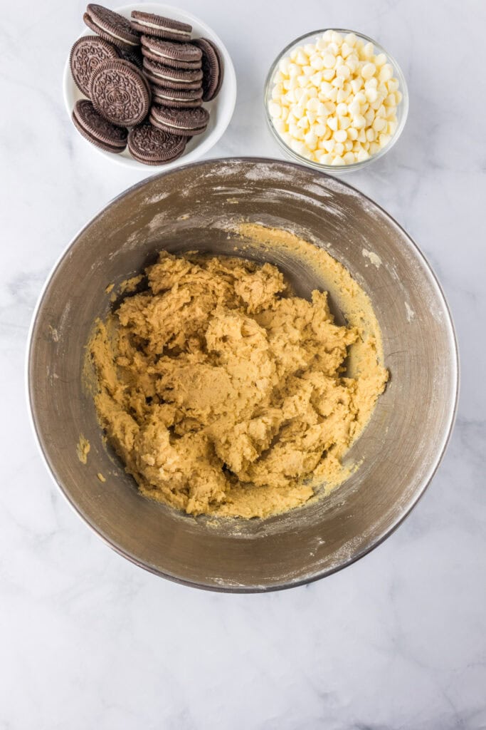 A metal mixing bowl with cookie dough for Oreo pudding cookies sits next to a bowl of white chocolate chips and a plate of chocolate sandwich cookies on a marble countertop.