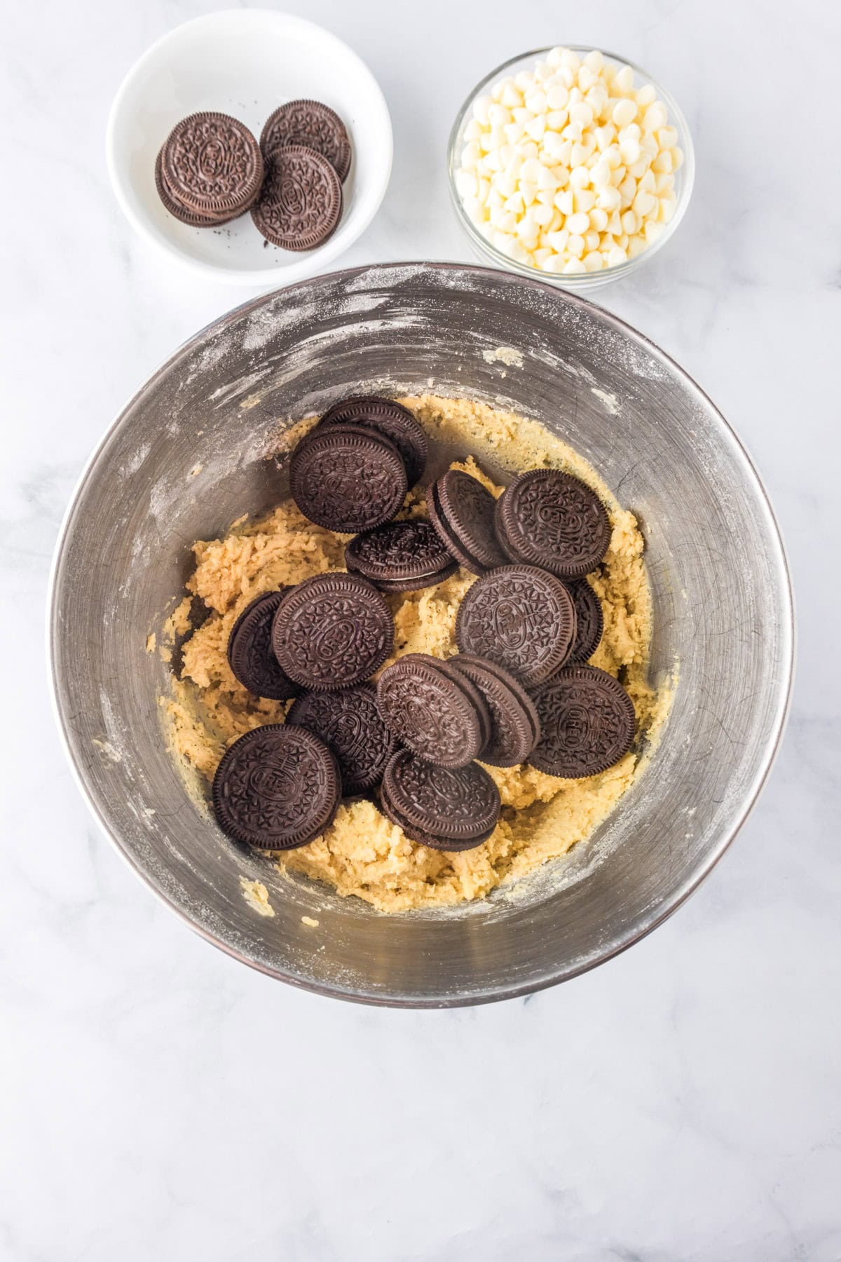 A mixing bowl with cookie dough and whole chocolate sandwich cookies for making Oreo pudding cookies, next to a bowl of white chocolate chips and a plate with more cookies on a marble countertop.