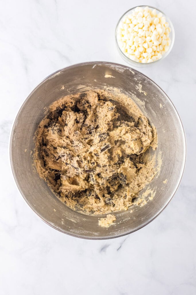A metal mixing bowl with cookie dough for Oreo pudding cookies sits on a white surface next to a small glass bowl filled with white chocolate chips.