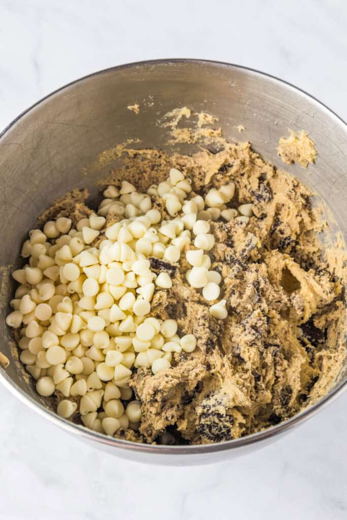 Stainless steel mixing bowl containing partially mixed oreo pudding cookies dough with white chocolate chips on top being mixed in from the side.