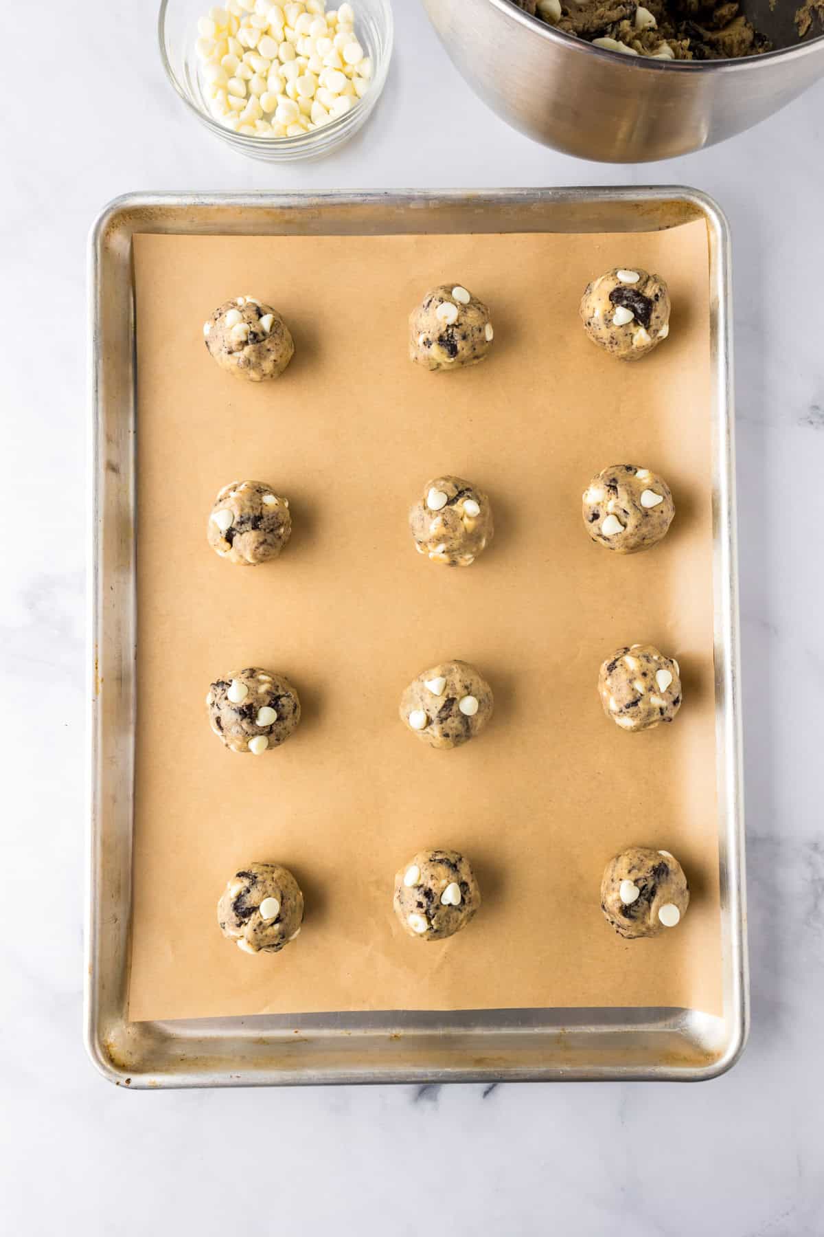 A baking sheet lined with parchment paper holds twelve balls of Oreo pudding cookie dough, ready to be baked. A bowl of white chocolate chips is visible in the background.