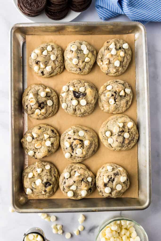 A baking tray lined with parchment paper holds twelve oreo pudding cookies topped with white chocolate chips. on pan after baking.