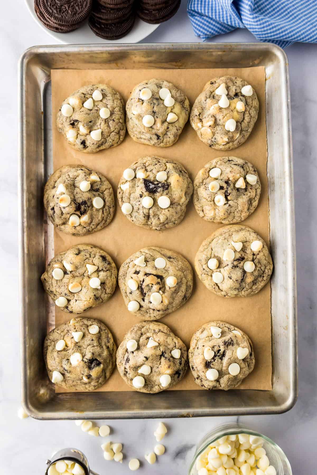 A baking tray lined with parchment paper holds twelve oreo pudding cookies topped with white chocolate chips. on pan after baking.