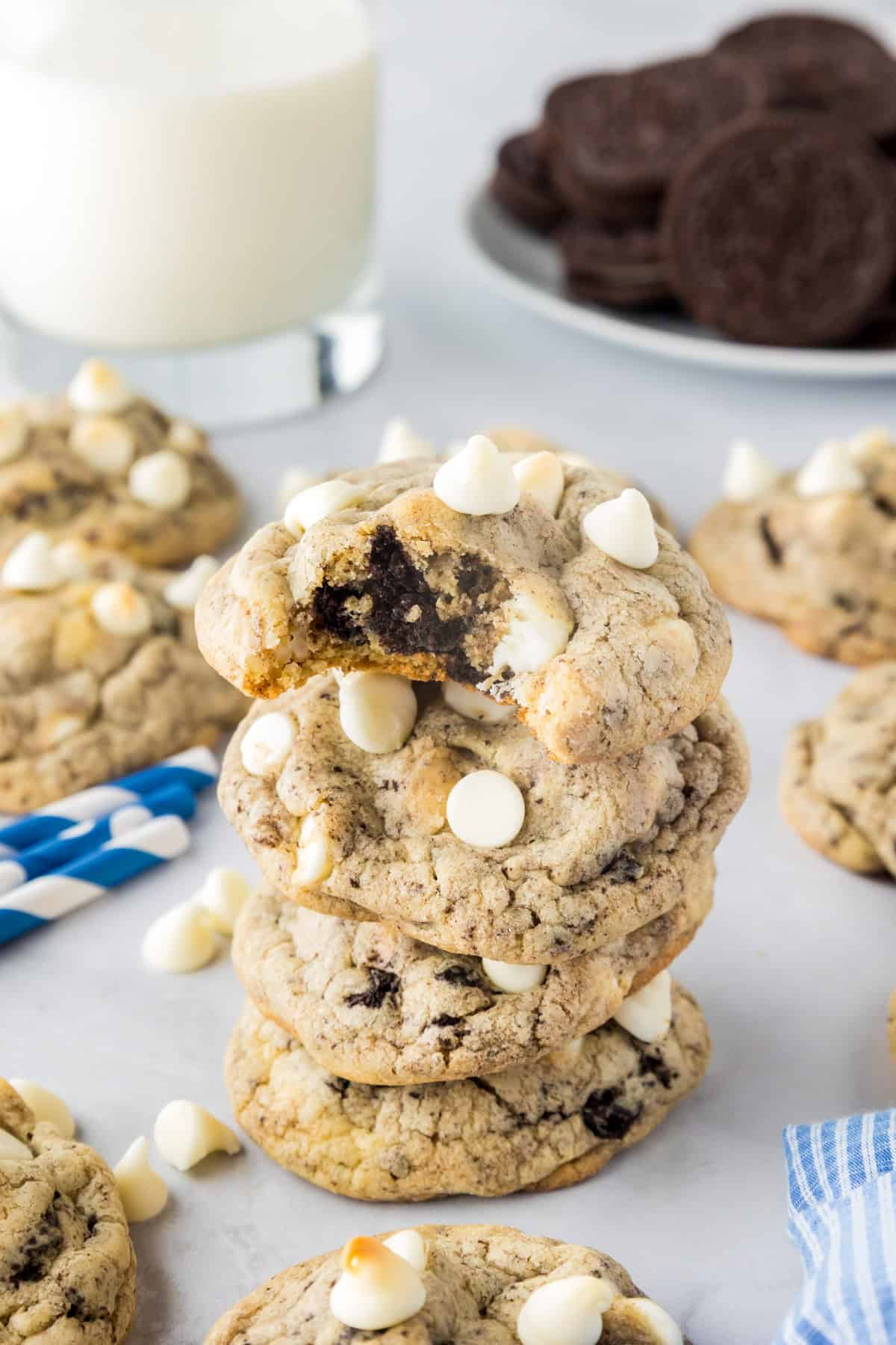 A stack of oreo pudding cookies with white chocolate chips and a bite taken from the top cookie, with a glass of milk and chocolate sandwich cookies in the background.