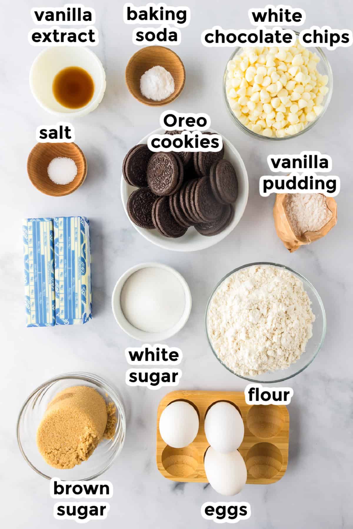 A variety of labeled baking ingredients on a marble surface, perfect for making Oreo pudding cookies—including eggs, sugars, butter, flour, white chocolate chips, Oreo cookies, vanilla pudding, vanilla extract, salt, and baking soda.