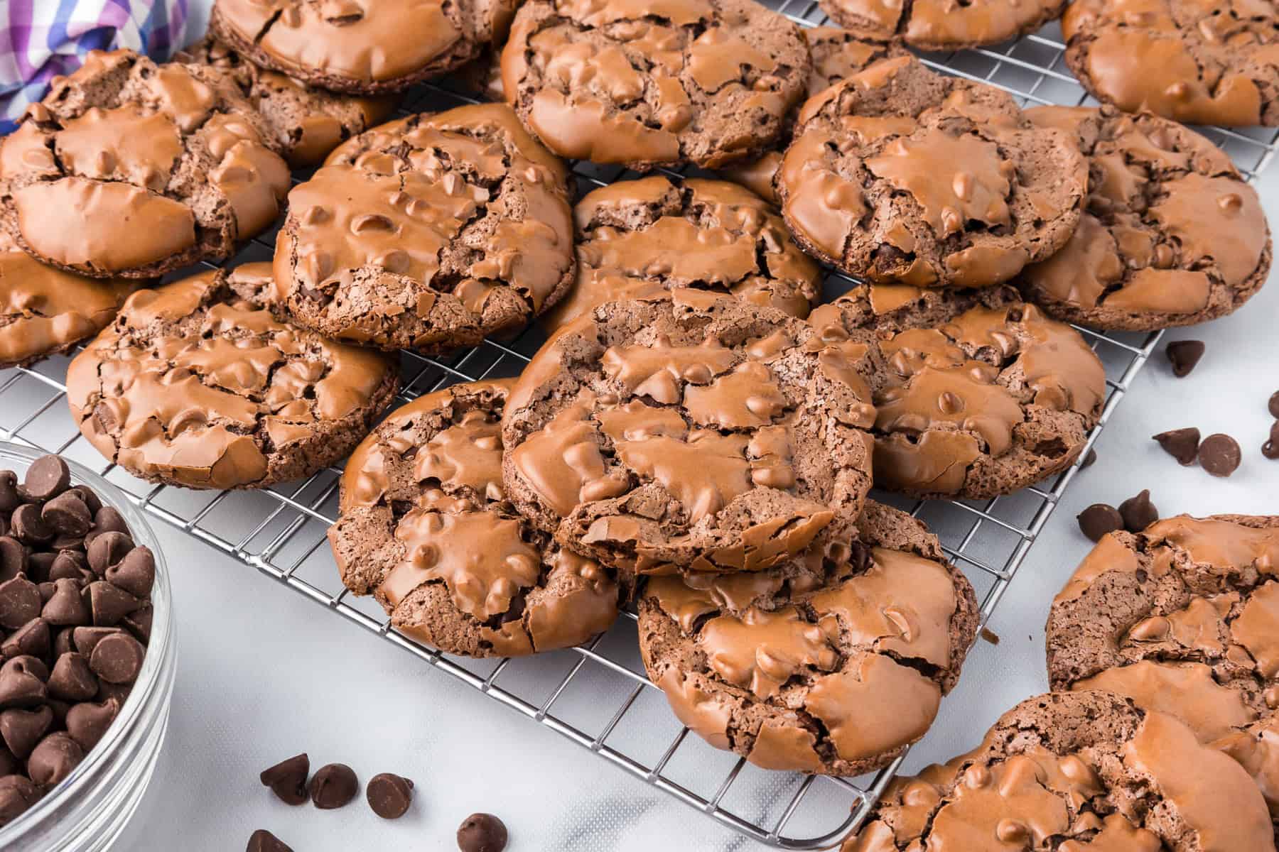 Flourless brownie cookies with a cracked surface cool on a wire rack, surrounded by a bowl of chocolate chips and scattered chips nearby.