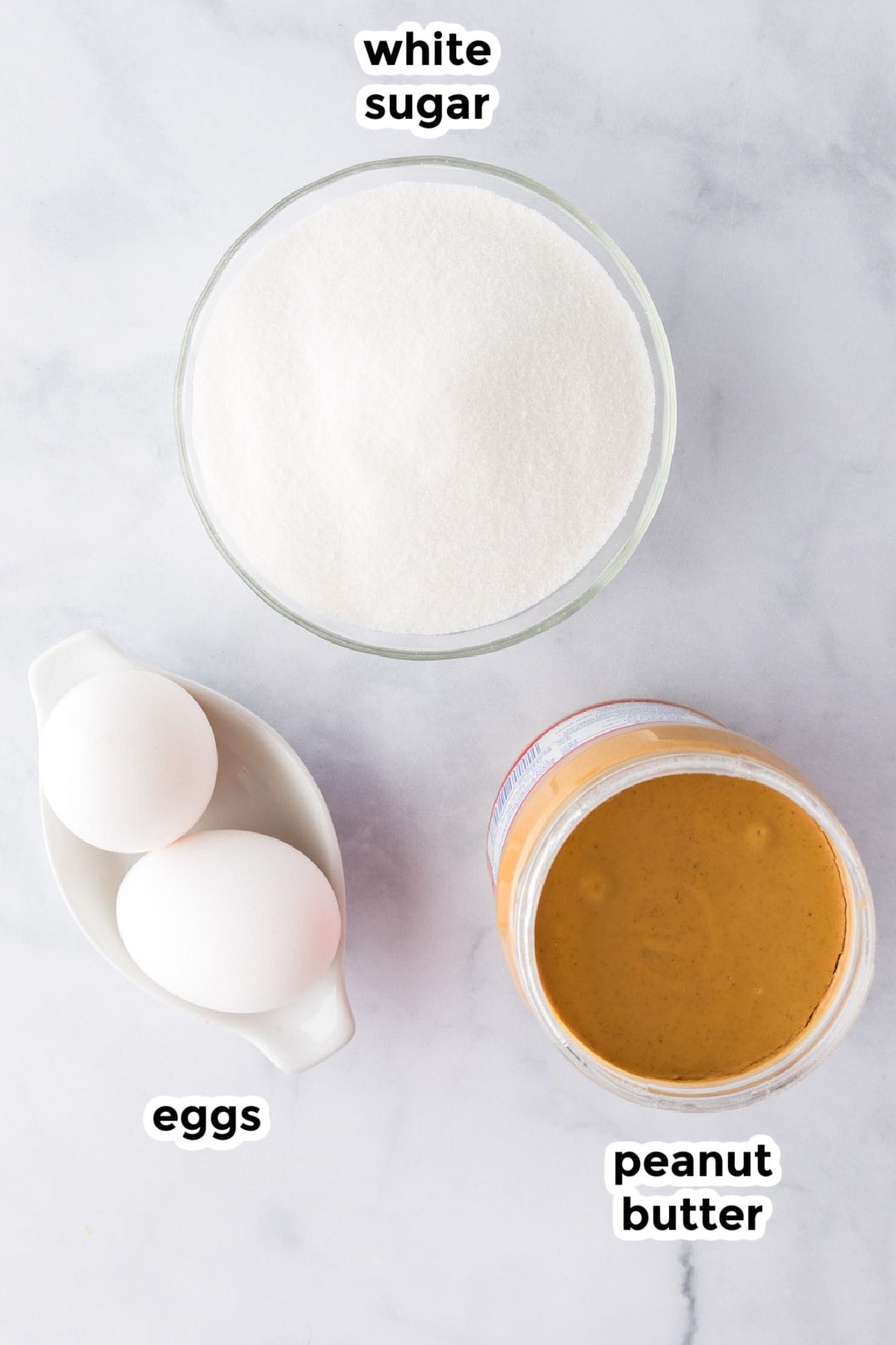 A bowl of white sugar, two eggs in a dish, and an open jar of peanut butter on a white surface—labeled as the simple ingredients for 3 ingredient peanut butter cookies.