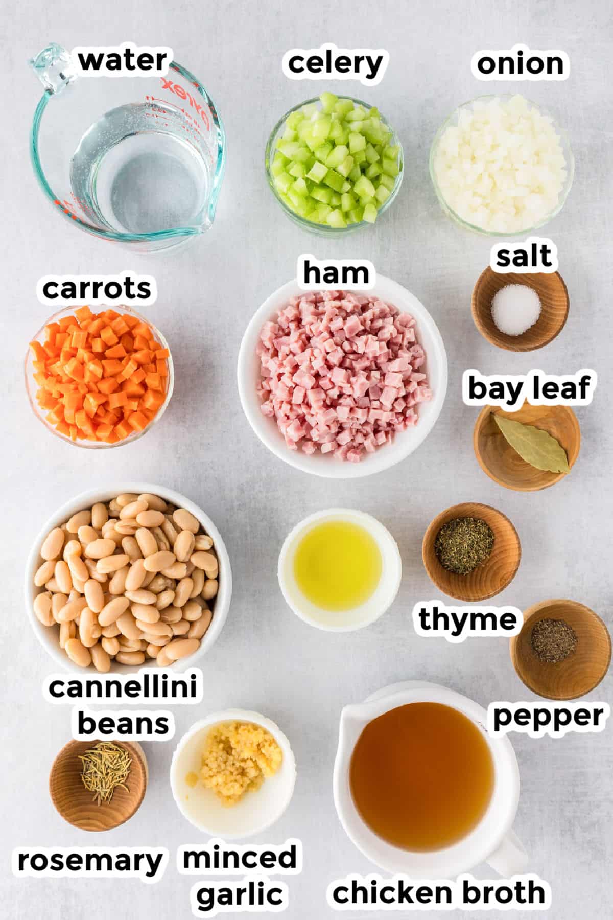 Ingredients for white bean and ham soup displayed in bowls and labeled: water, celery, onion, salt, bay leaf, ham, carrots, cannellini beans, olive oil, thyme, pepper, rosemary, minced garlic, chicken broth.