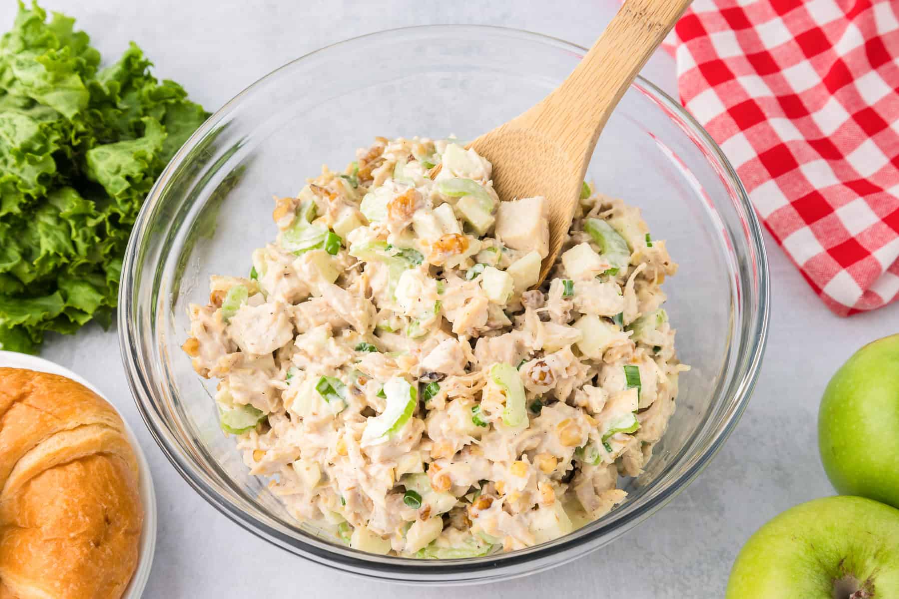 A glass bowl of apple walnut chicken salad with chopped celery, walnuts, and herbs mixed with mayonnaise sits on a counter with a wooden spoon. Nearby are lettuce, a roll, a red-checkered cloth, and green apples.