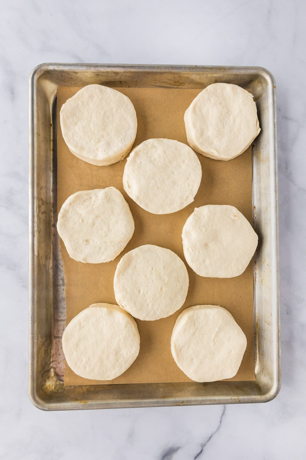 Eight unbaked biscuit rounds for Biscuit Chicken Pot Pie are arranged on a parchment-lined baking sheet, viewed from above on a marble surface.