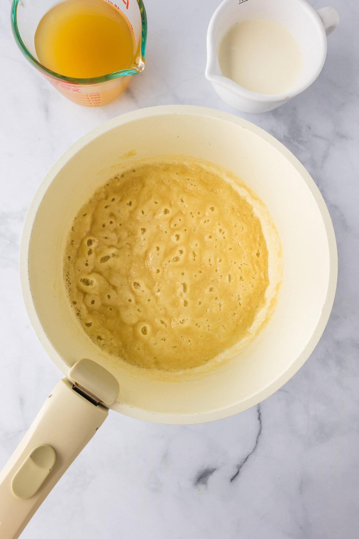 A white saucepan on a marble surface contains a light brown bubbling sauce,just the start of a comforting Biscuit Chicken Pot Pie. Nearby are a measuring cup of broth and a small pitcher of cream, ready to make the dish extra creamy.