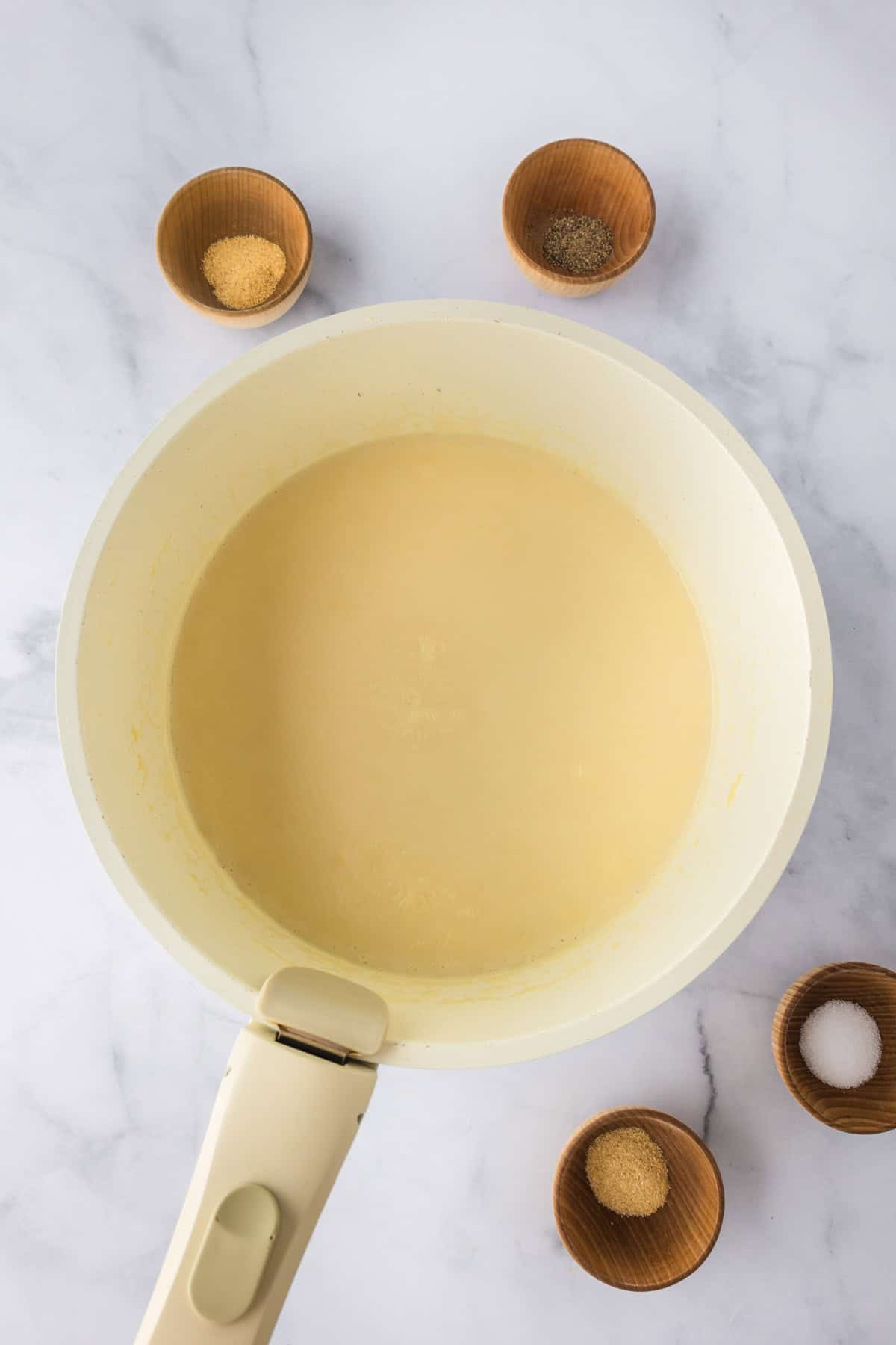A white saucepan with a creamy Biscuit Chicken Pot Pie filling inside, surrounded by four small wooden bowls containing various spices, on a white marble surface.