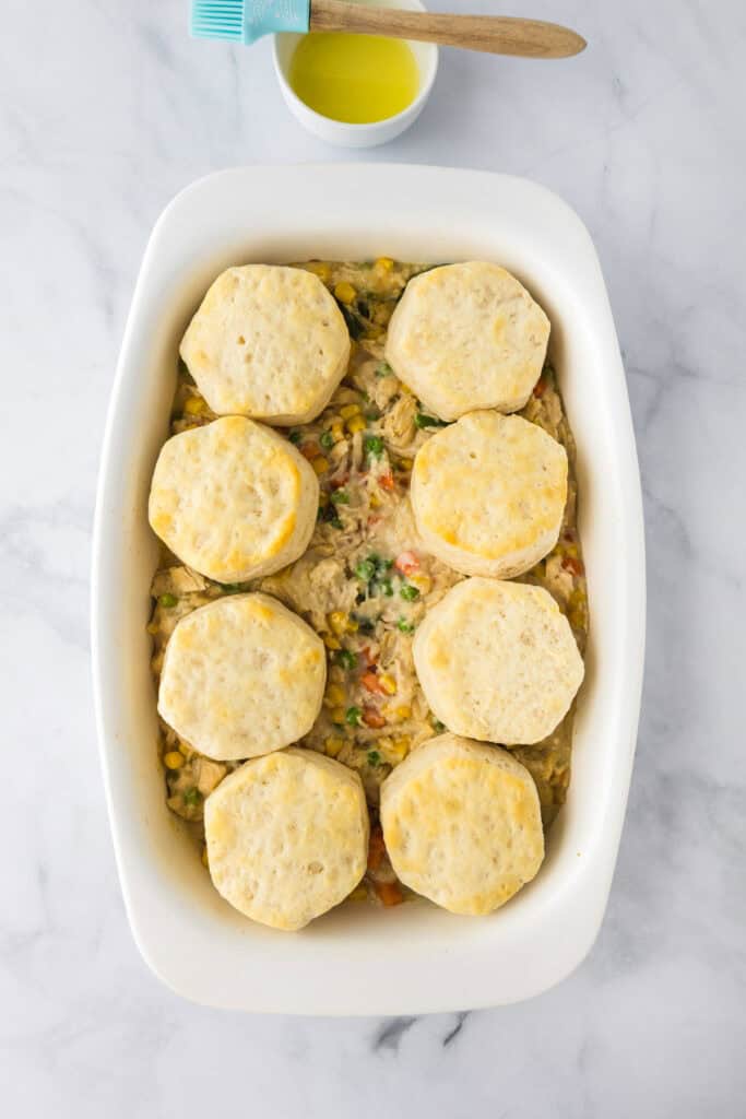 A white baking dish holds a creamy vegetable mixture for Biscuit Chicken Pot Pie, topped with eight round, unbaked biscuits; a small bowl with a pastry brush sits nearby.