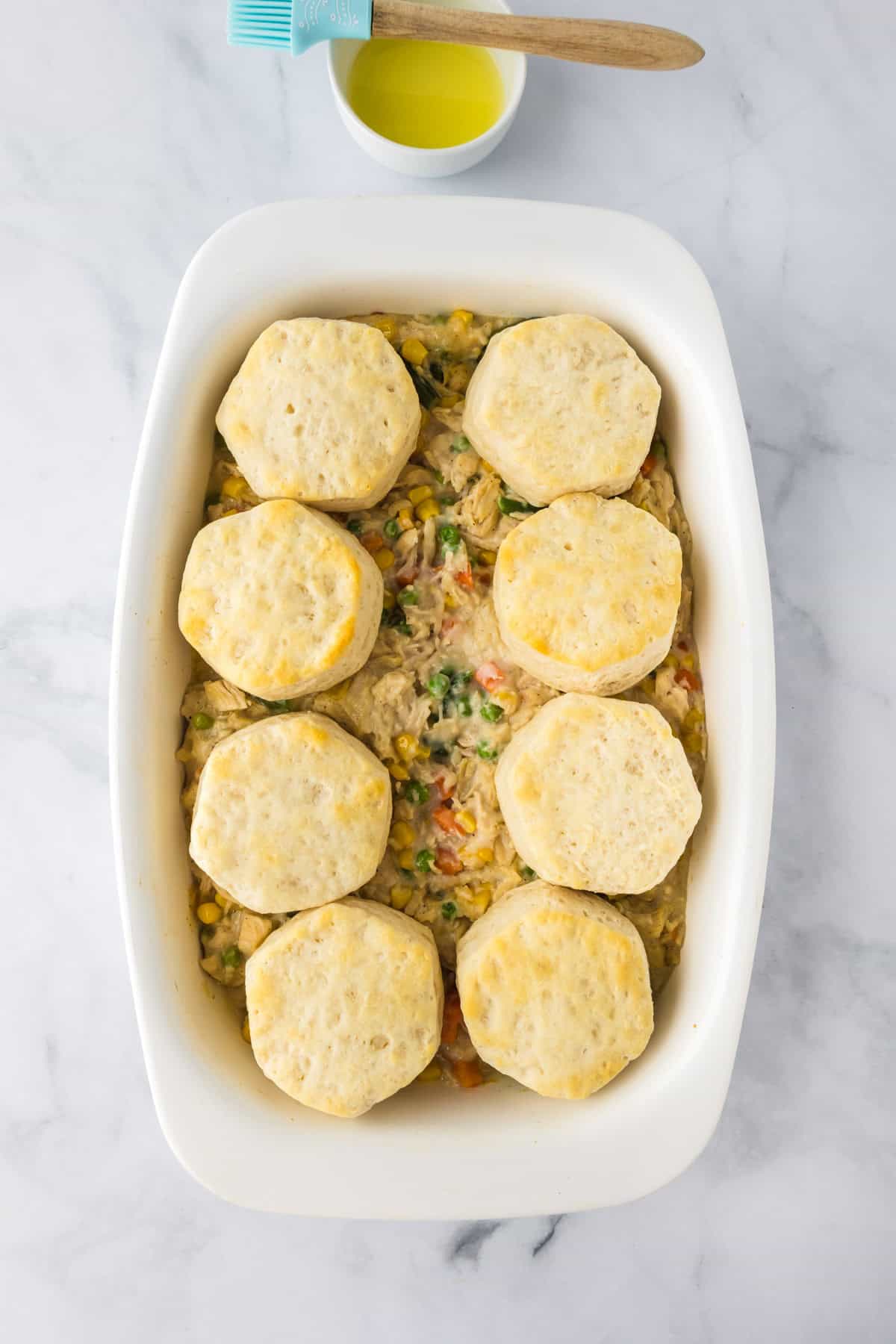 A white baking dish holds a creamy vegetable mixture for Biscuit Chicken Pot Pie, topped with eight round, unbaked biscuits; a small bowl with a pastry brush sits nearby.