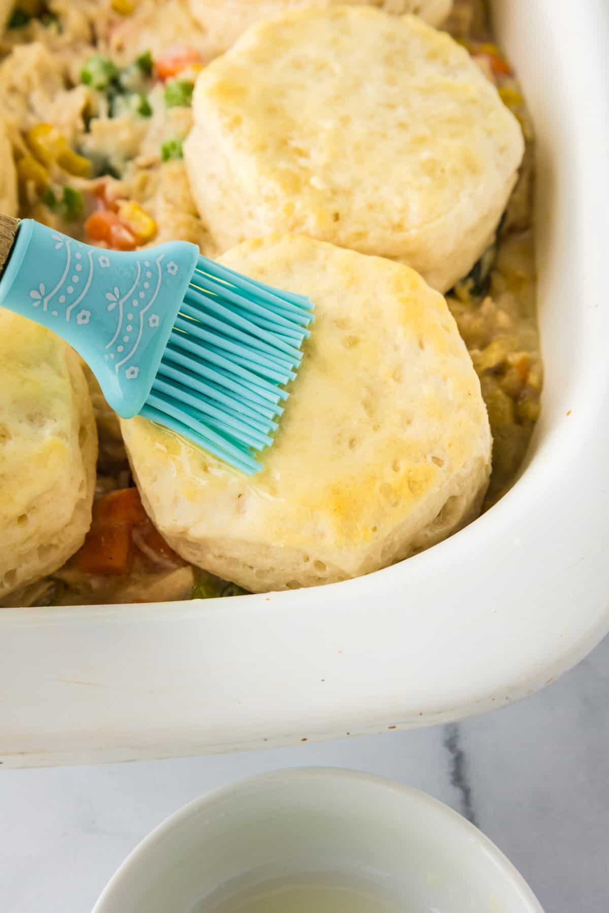 A silicone brush spreads butter or egg wash on top of uncooked biscuits layered over a Biscuit Chicken Pot Pie in a white baking dish.