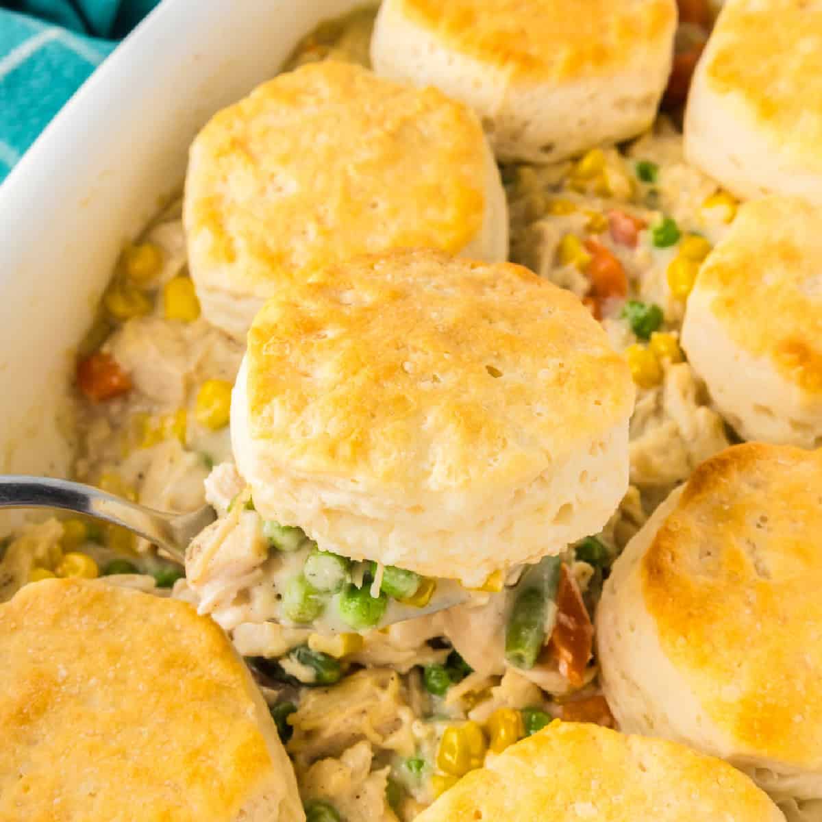 A casserole dish filled with creamy biscuit chicken pot pie and mixed vegetables, topped with golden brown biscuits. A spoon is serving a portion.