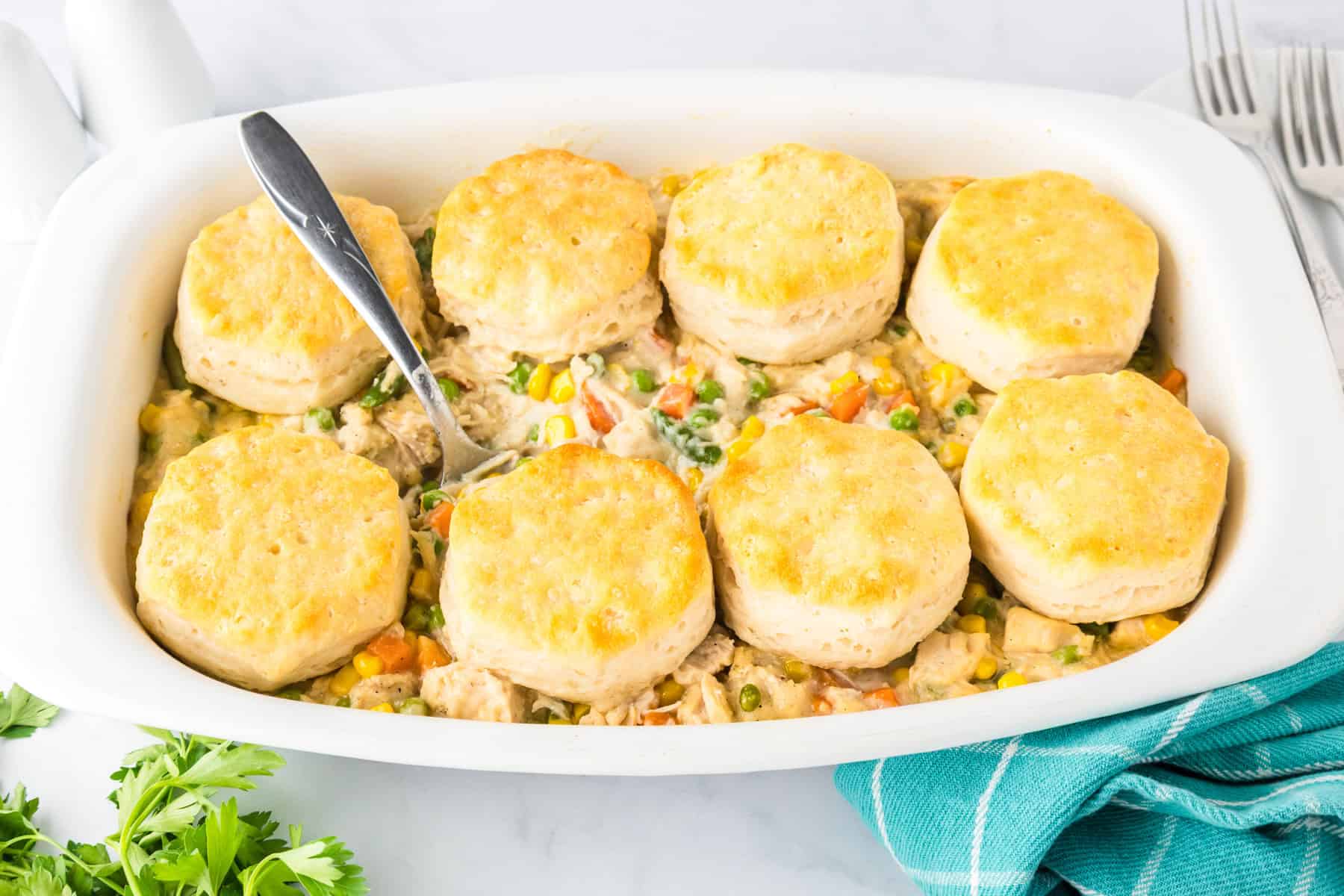 A white casserole dish filled with biscuit chicken pot pie features tender chicken and vegetables in a creamy filling, topped with eight golden biscuits and a serving spoon inside.