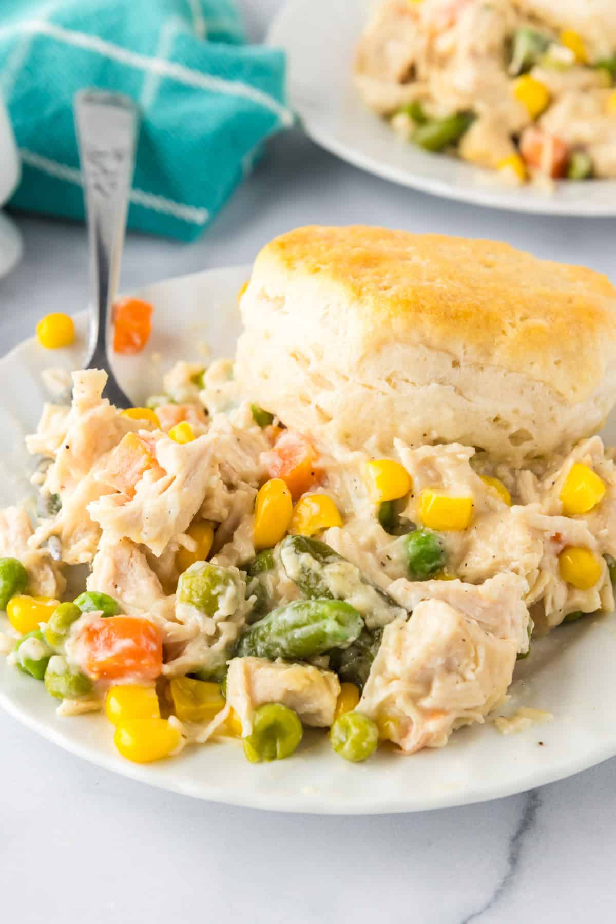 A plate of biscuit chicken pot pie casserole topped with a golden biscuit, featuring chunks of chicken, carrots, peas, green beans, and corn in a creamy sauce.