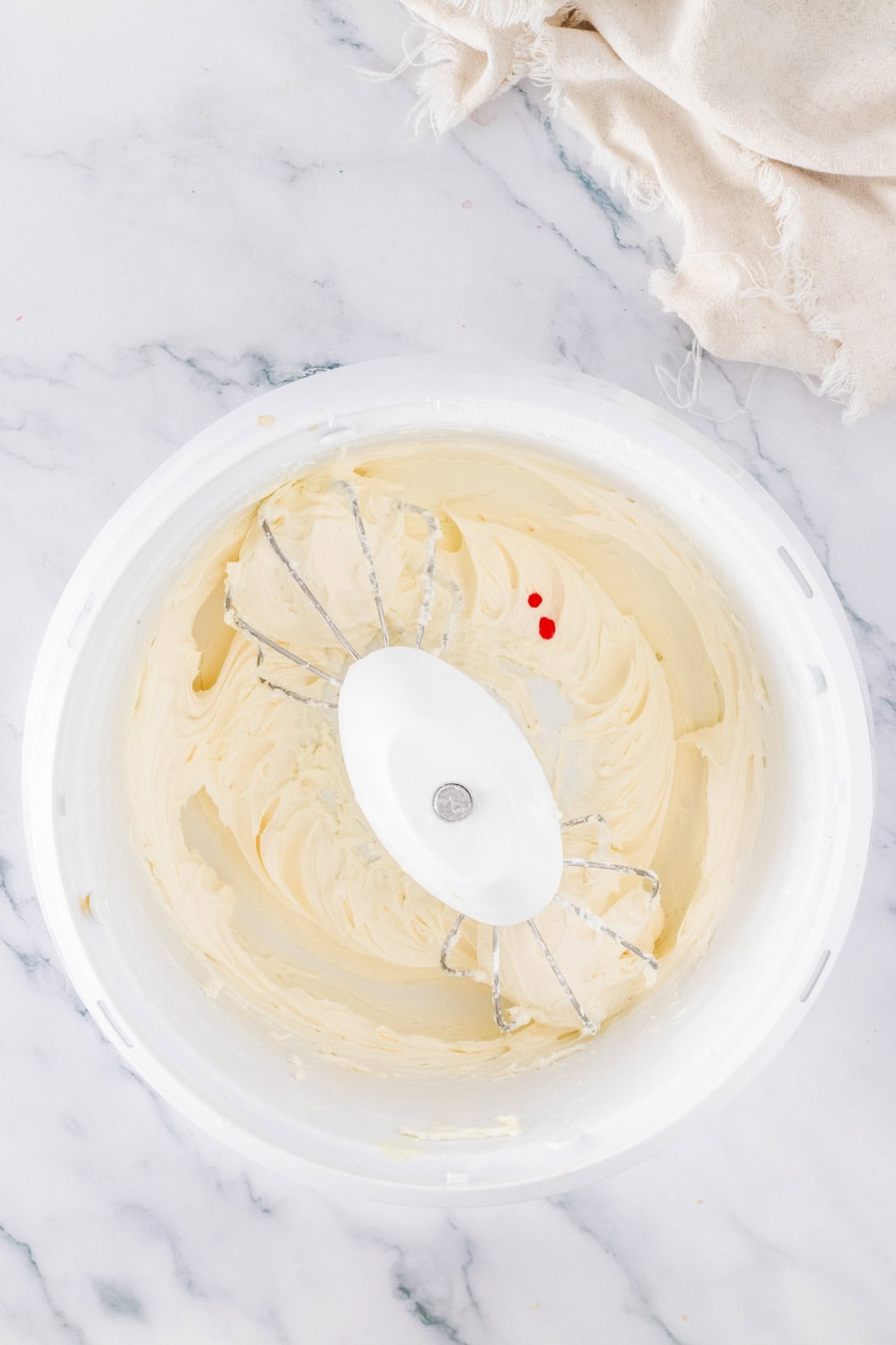 A white mixing bowl with creamed batter, two drops of red food coloring, and two metal beaters on a marble surface—perfect for making Cadbury Mini Egg Cookie Bars—with a beige cloth nearby.