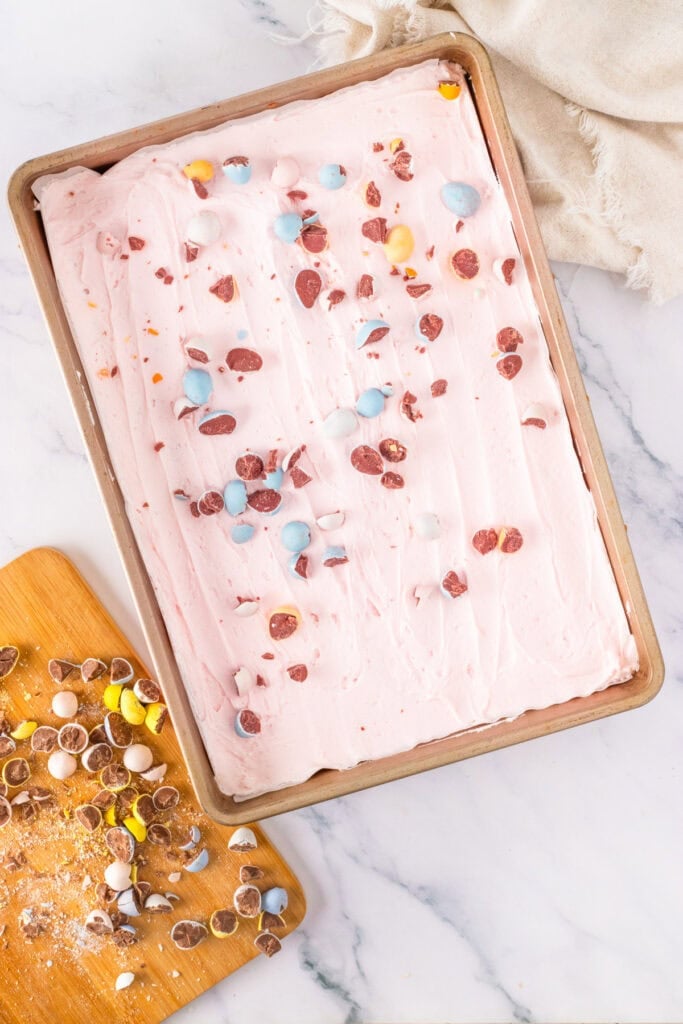 A sheet cake with pink frosting, topped with crushed chocolate eggs, sits on a marble surface next to a cutting board with more Cadbury Mini Egg Cookie Bars and crushed chocolate eggs.