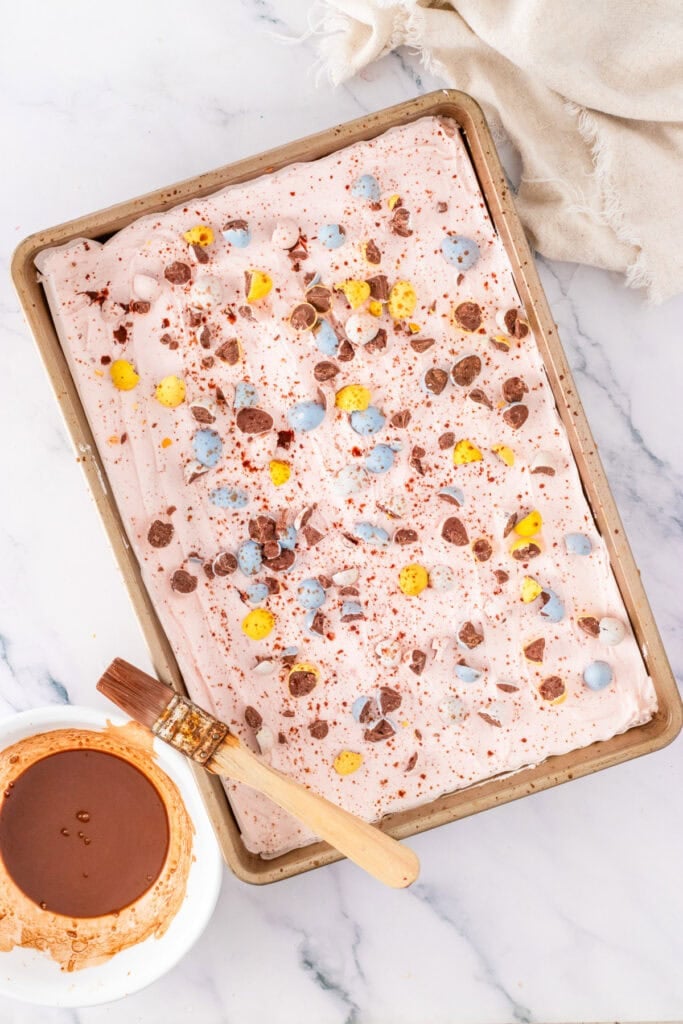 A rectangular pan filled with pink Cadbury Mini Egg Cookie Bars, topped with crushed candy eggs and chocolate, sits beside a dish of liquidy chocolate and a pastry brush on a marble surface.