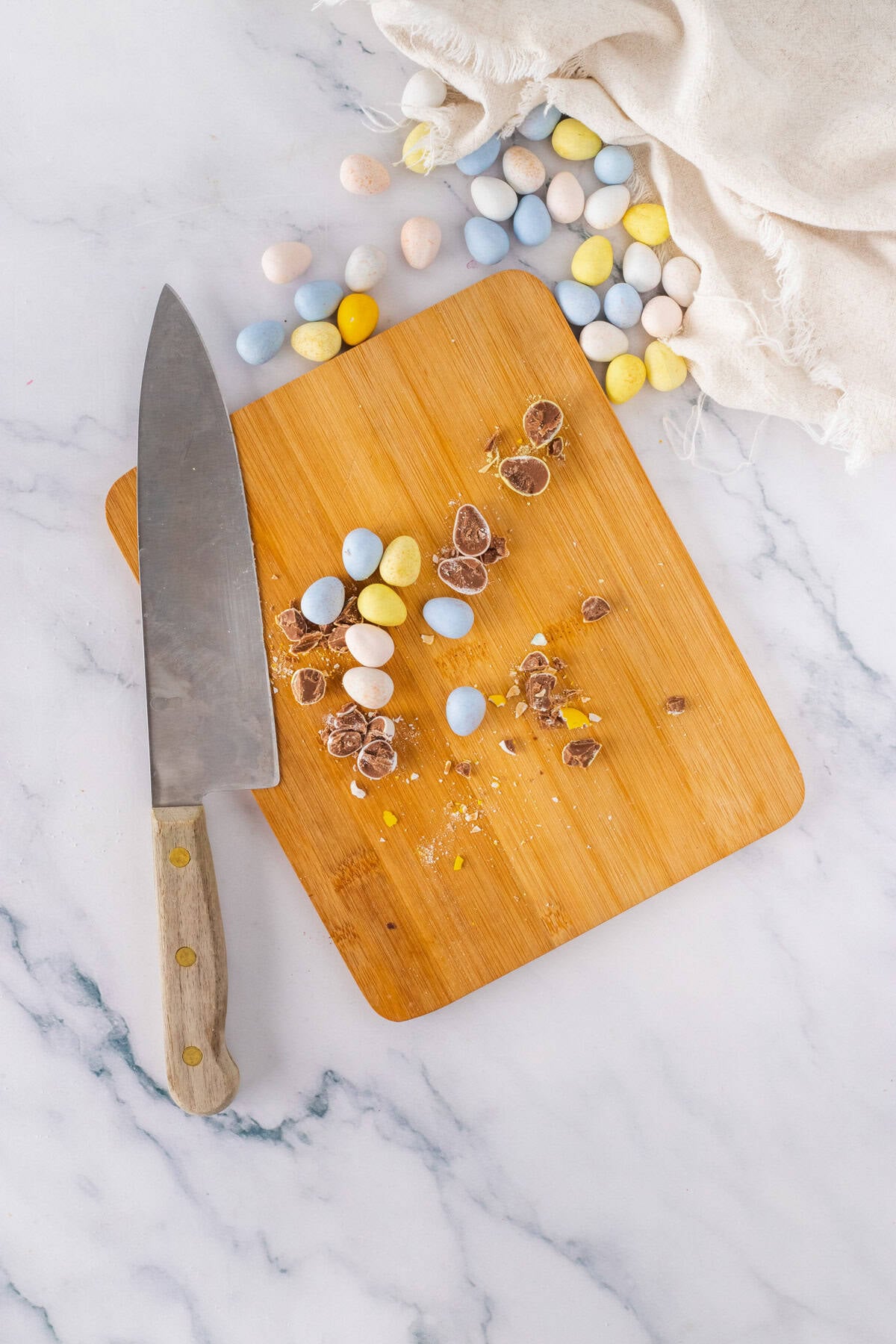 A wooden cutting board with chocolate eggs, some cut open, next to a large knife on a marble surface for Cadbury Mini Egg Cookie Bars.