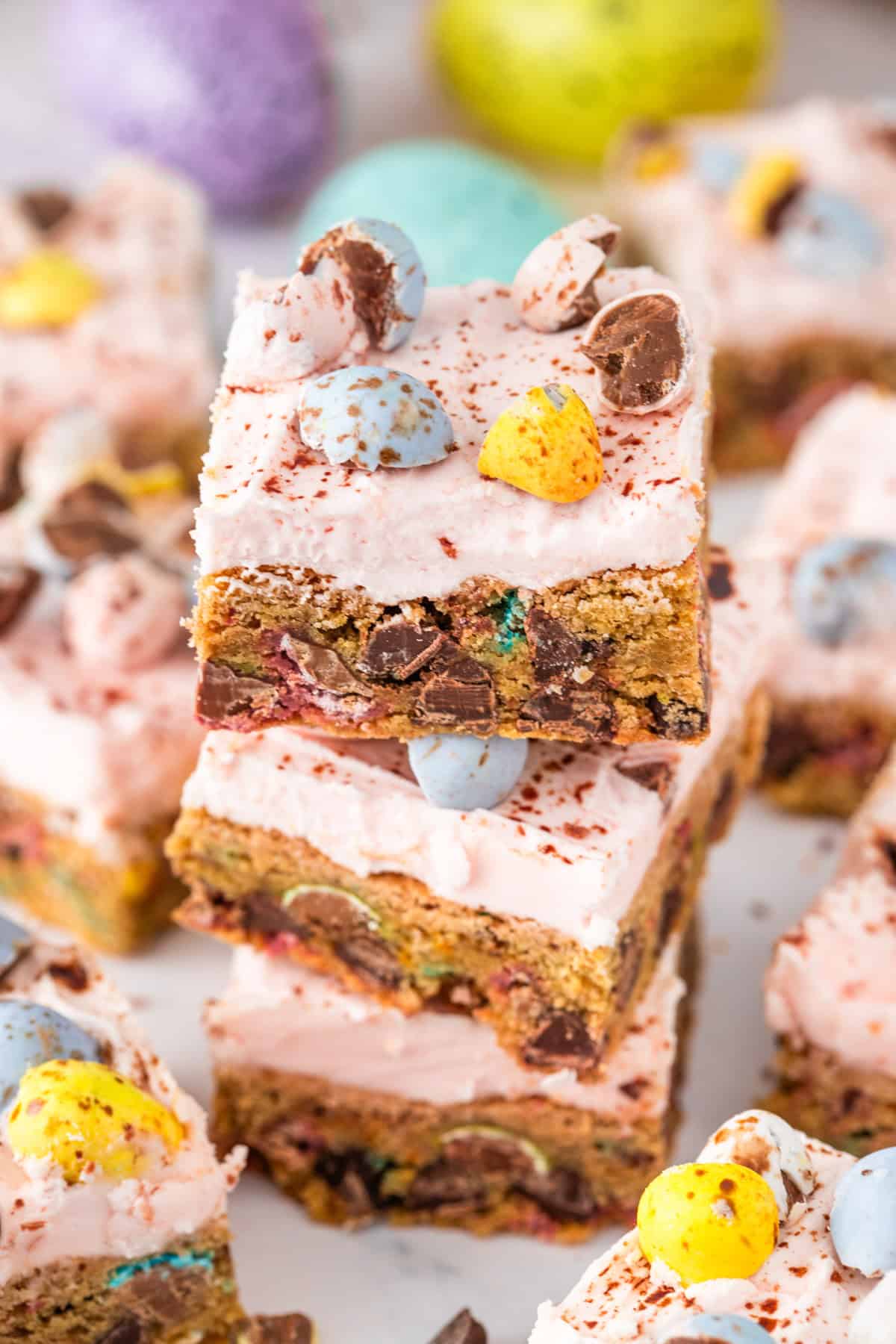 Three Cadbury Mini Egg Cookie Bars are stacked, each topped with halved chocolate eggs and sprinkles, with more bars and pastel candies in the background.