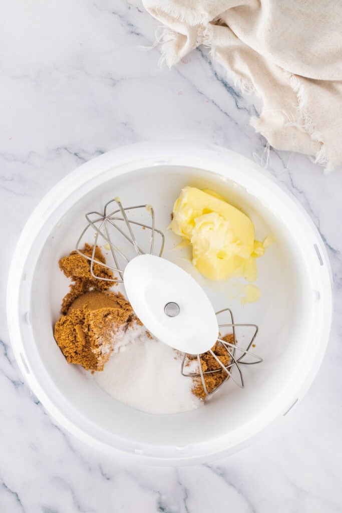 A mixing bowl with brown sugar, white sugar, and butter sits ready for Cadbury Mini Egg Cookie Bars, with a stand mixer attachment on a marble countertop/
