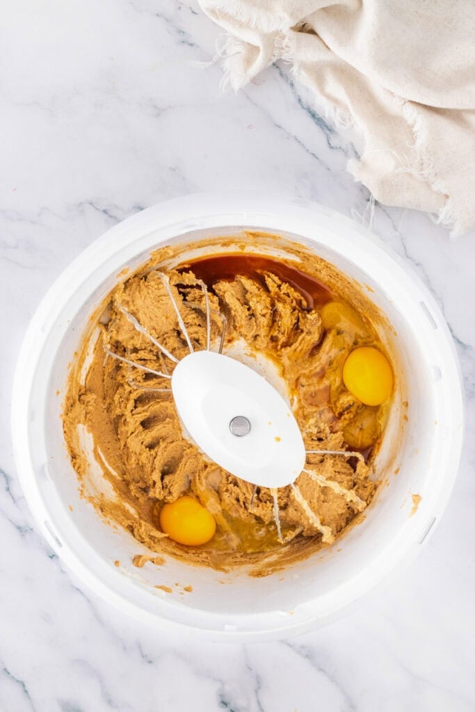 A mixing bowl with Cadbury Mini Egg Cookie Bars wet ingredients, two raw eggs, and vanilla extract partially blended together, placed on a marble countertop next to a beige cloth.