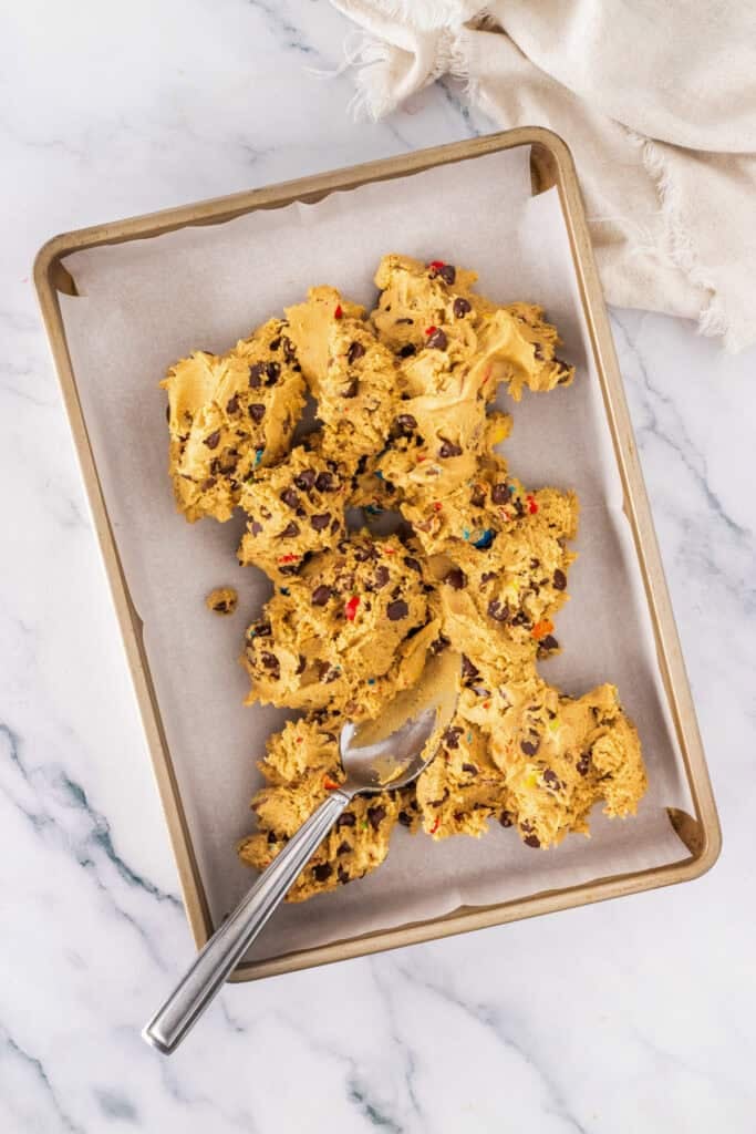 A baking sheet lined with parchment paper holds scoops of Cadbury Mini Egg Cookie Bars dough, dotted with chocolate chips and colorful candies. A metal spoon rests on the sheet.