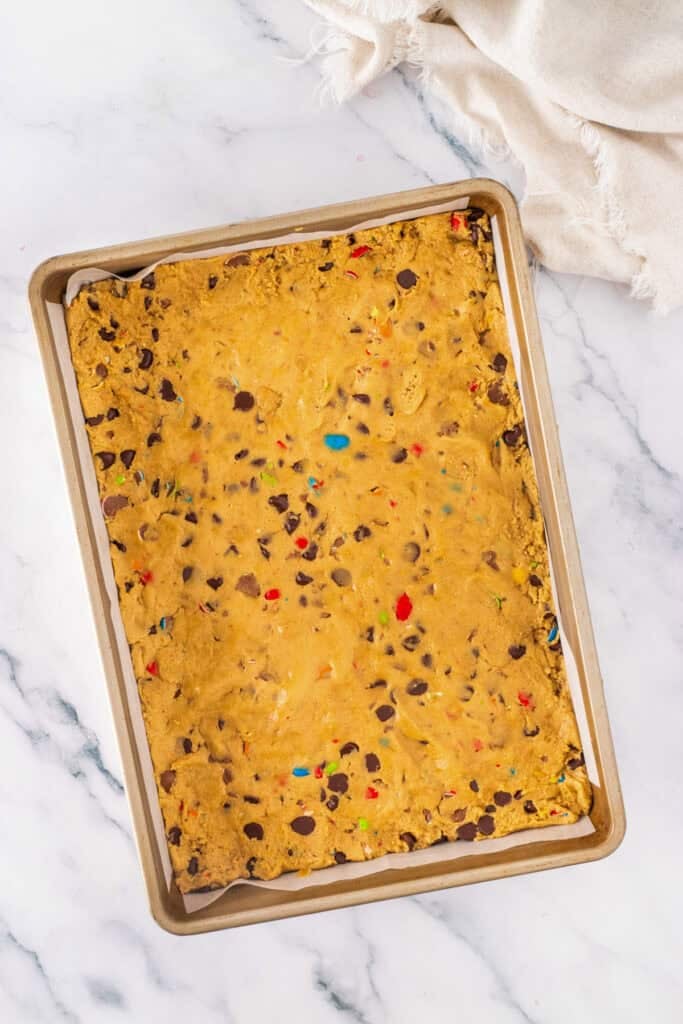 A rectangular pan filled with unbaked Cadbury Mini Egg Cookie Bars, their dough studded with chocolate chips and colorful candy pieces, set on a marble surface.
