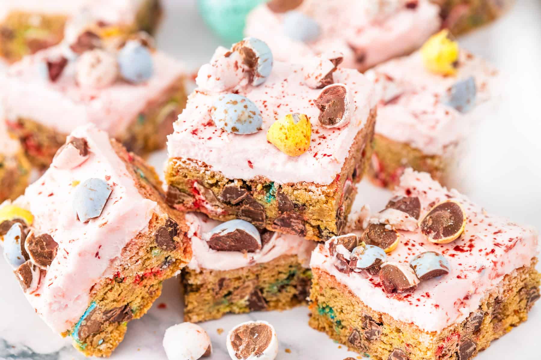 Several Cadbury Mini Egg Cookie Bars, topped with pink frosting and chopped candy-coated chocolate eggs, are arranged on a white surface.