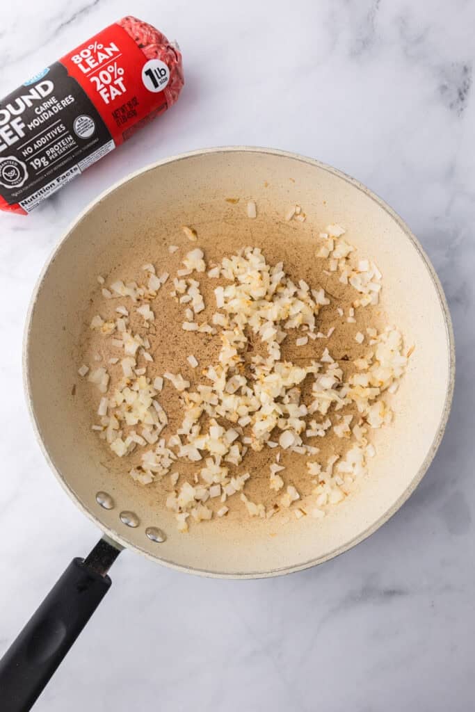 Chopped onions and garlic are sautéing in a light-colored skillet, with a package of ground beef on the countertop nearby—perfect beginnings for tasty cheeseburger sliders.