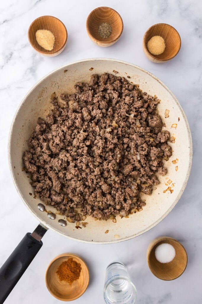 Cooked ground beef with onions in a skillet, perfect for making cheeseburger sliders, surrounded by small bowls of seasoning, spices, and a glass of water on a marble countertop.