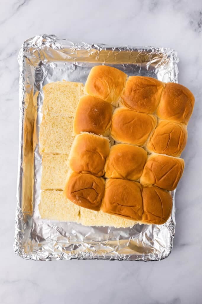 A foil-lined baking sheet holds a group of soft dinner rolls, half of which have been sliced horizontally, perfect for assembling delicious cheeseburger sliders.