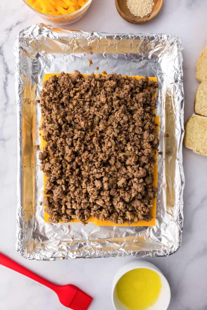 A foil-lined baking sheet features cheeseburger sliders, with layers of cheese slices and cooked ground beef, surrounded by shredded cheese, sesame seeds, bread, and a bowl of melted butter.