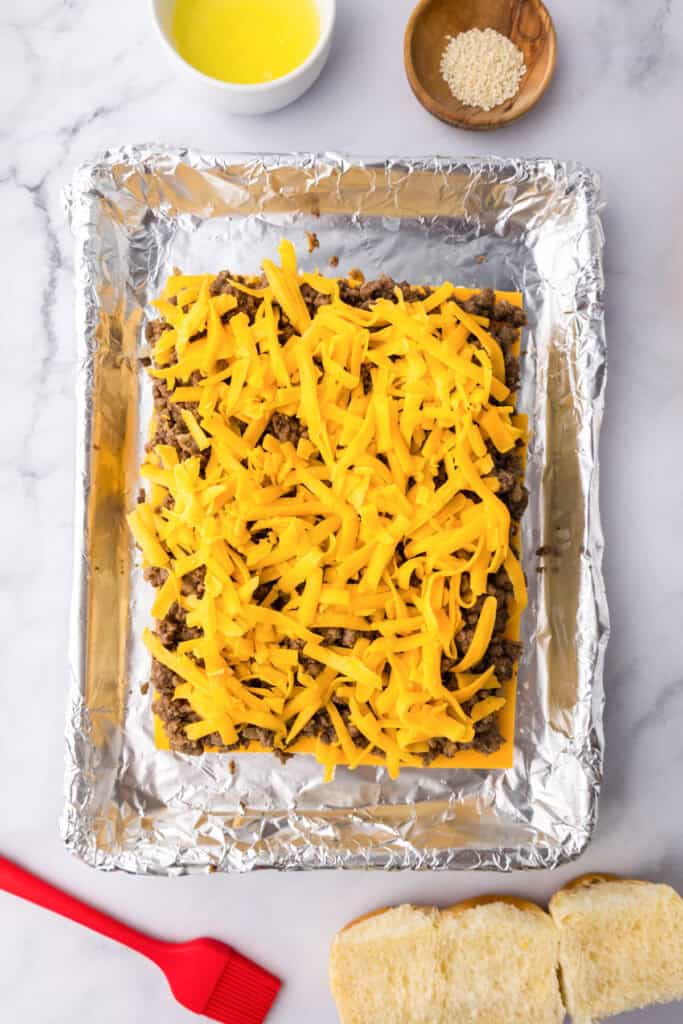 A foil-lined baking pan filled with a layer of ground beef and shredded cheddar cheese for cheeseburger sliders sits on a marble surface, surrounded by butter, sesame seeds, a red brush, and sliced bread.