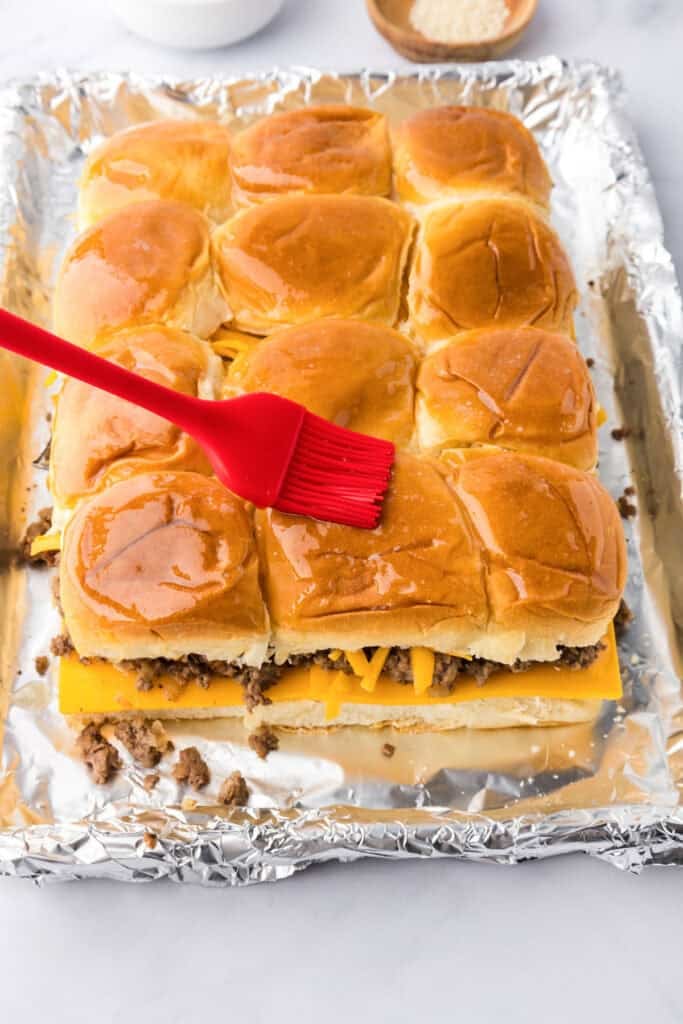 A red brush spreads melted butter on top of assembled cheeseburger sliders filled with ground beef and cheese, placed on a foil-lined baking sheet.