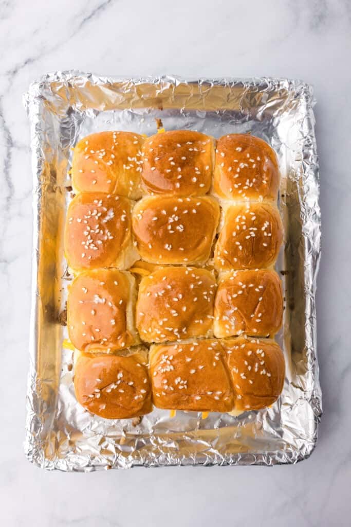 A tray of twelve golden brown dinner rolls, perfect for making cheeseburger sliders, topped with coarse salt and arranged on a foil-lined baking sheet.