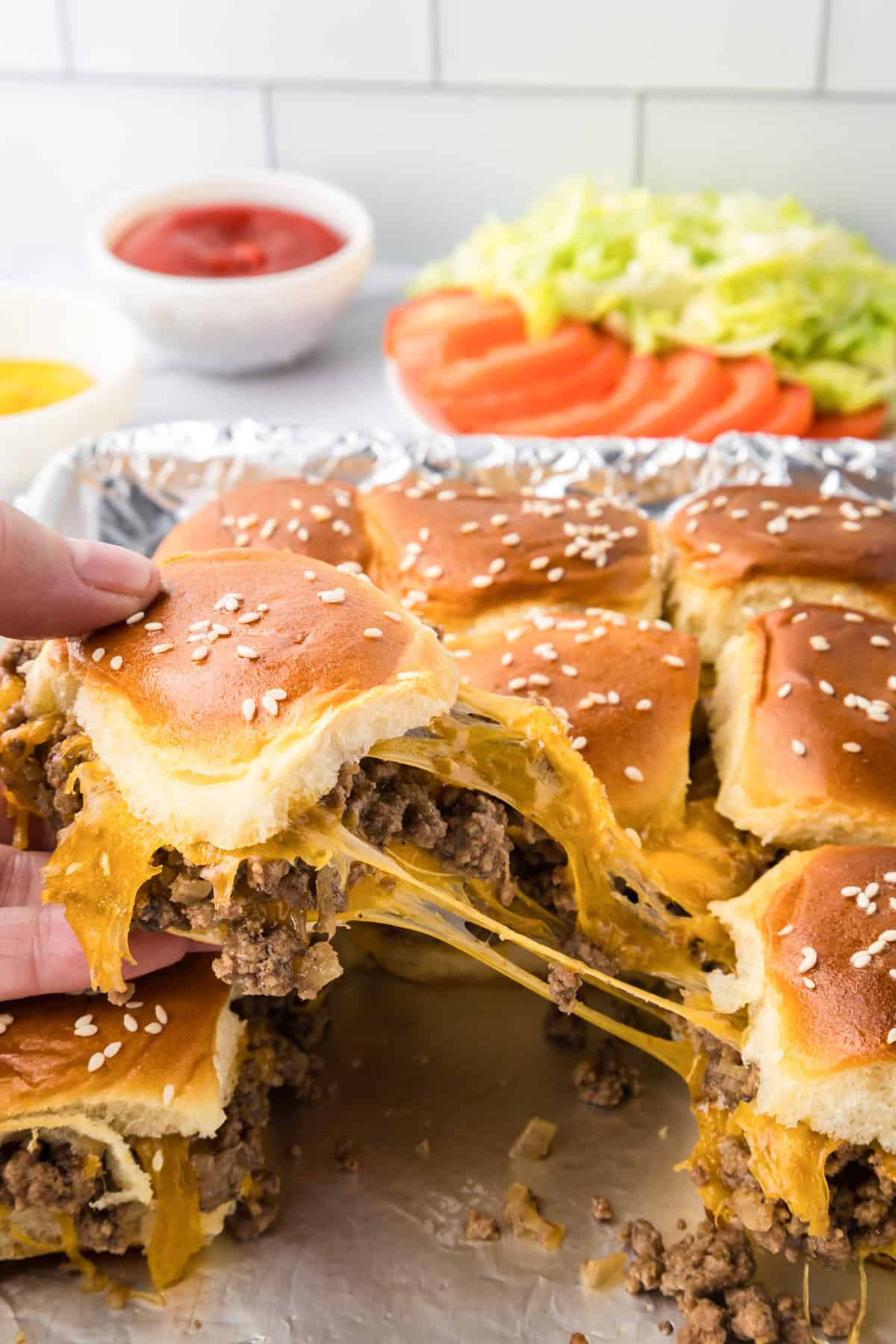 A hand lifts a cheesy ground beef cheeseburger slider with a sesame seed bun from a tray; other cheeseburger sliders, shredded lettuce, tomato slices, and sauces are in the background.