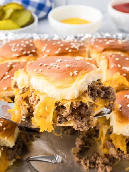 A metal spatula lifts a cheesy ground beef cheeseburger slider on a sesame bun from a tray of similar sliders, with pickles and condiments in the background.