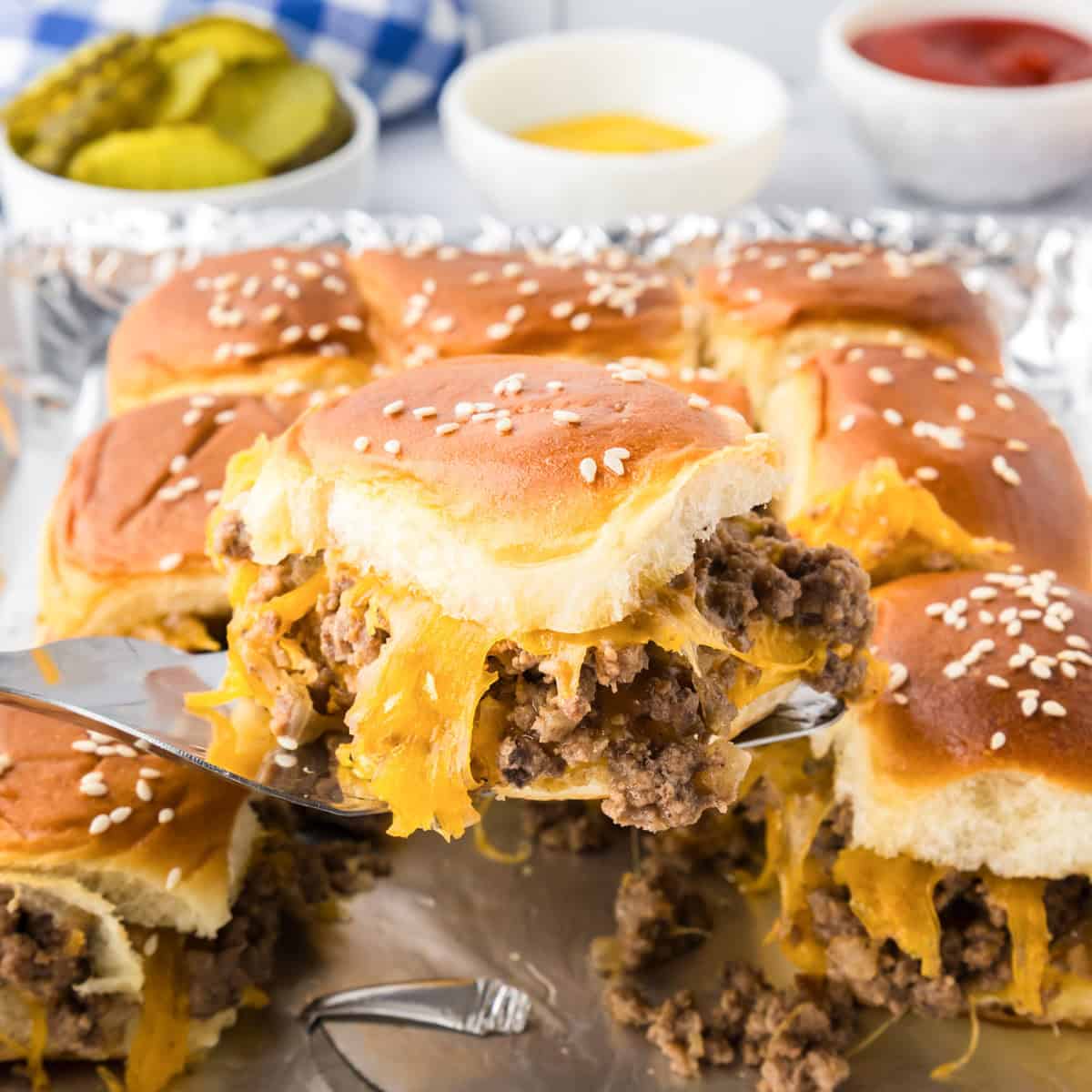 A metal spatula lifts a cheesy ground beef cheeseburger slider on a sesame bun from a tray of similar sliders, with pickles and condiments in the background.