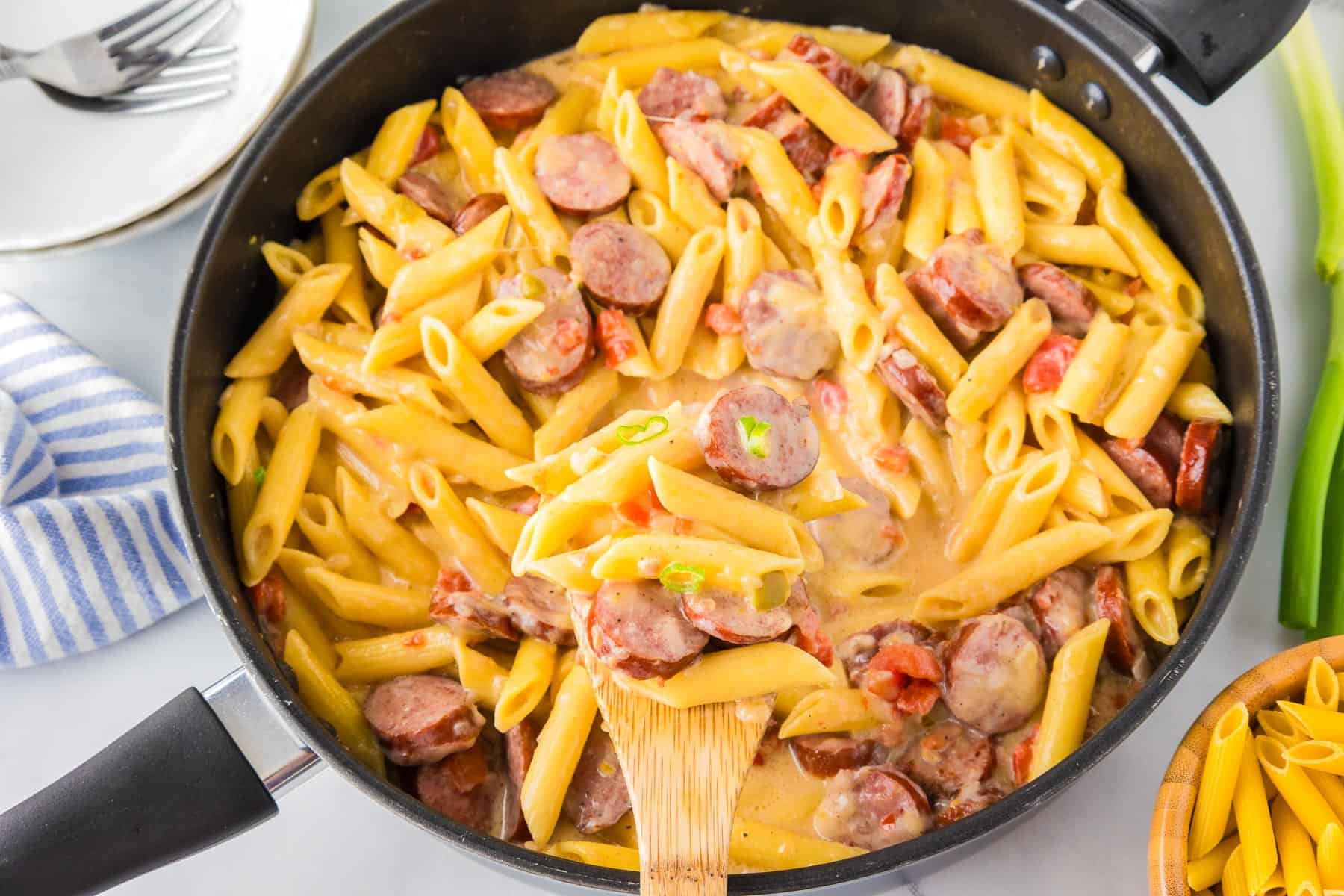 A skillet filled with creamy Cheesy Smoked Sausage Pasta—penne, sliced sausage, and diced tomatoes—all being stirred together with a wooden spoon.