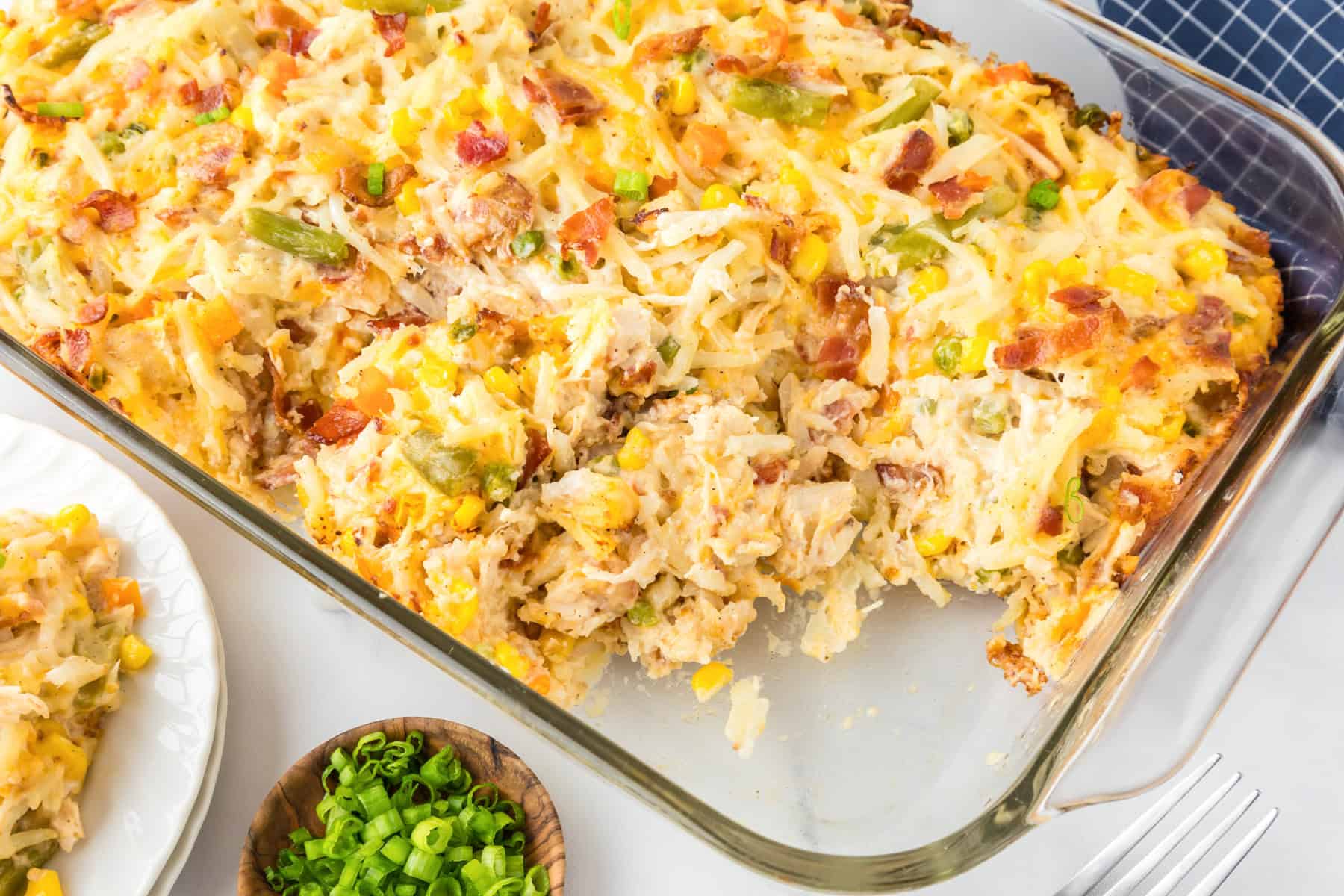 A hearty chicken hashbrown casserole with shredded cheese, corn, green beans, and bacon is baked in a glass dish with a portion scooped out. A small bowl of chopped green onions sits nearby.