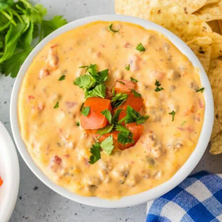A bowl of crock pot Rotel dip topped with chopped tomatoes and cilantro, surrounded by tortilla chips, diced tomatoes, and cilantro on a gray surface.
