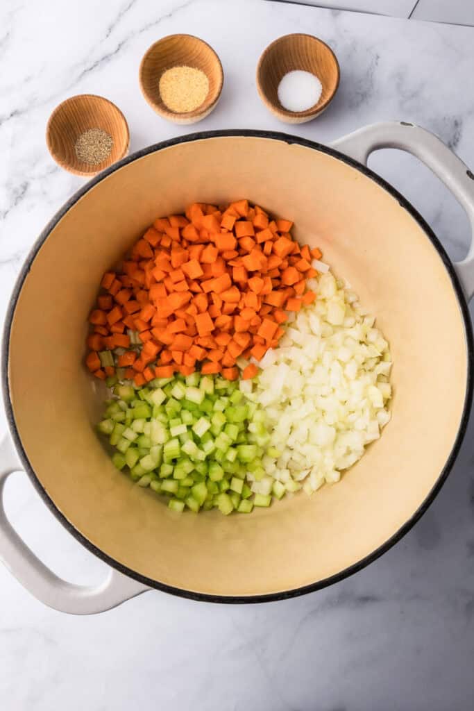 A large pot with diced carrots, celery, and onions ready for minestrone soup; three small bowls containing spices and salt sit above the pot on a marble countertop.